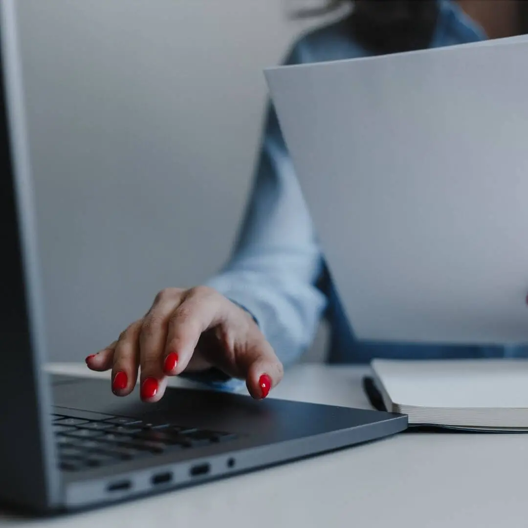 Mãos de uma profissional operando um notebook e manuseando papéis, simbolizando a organização técnica de documentos e ementas acadêmicas.